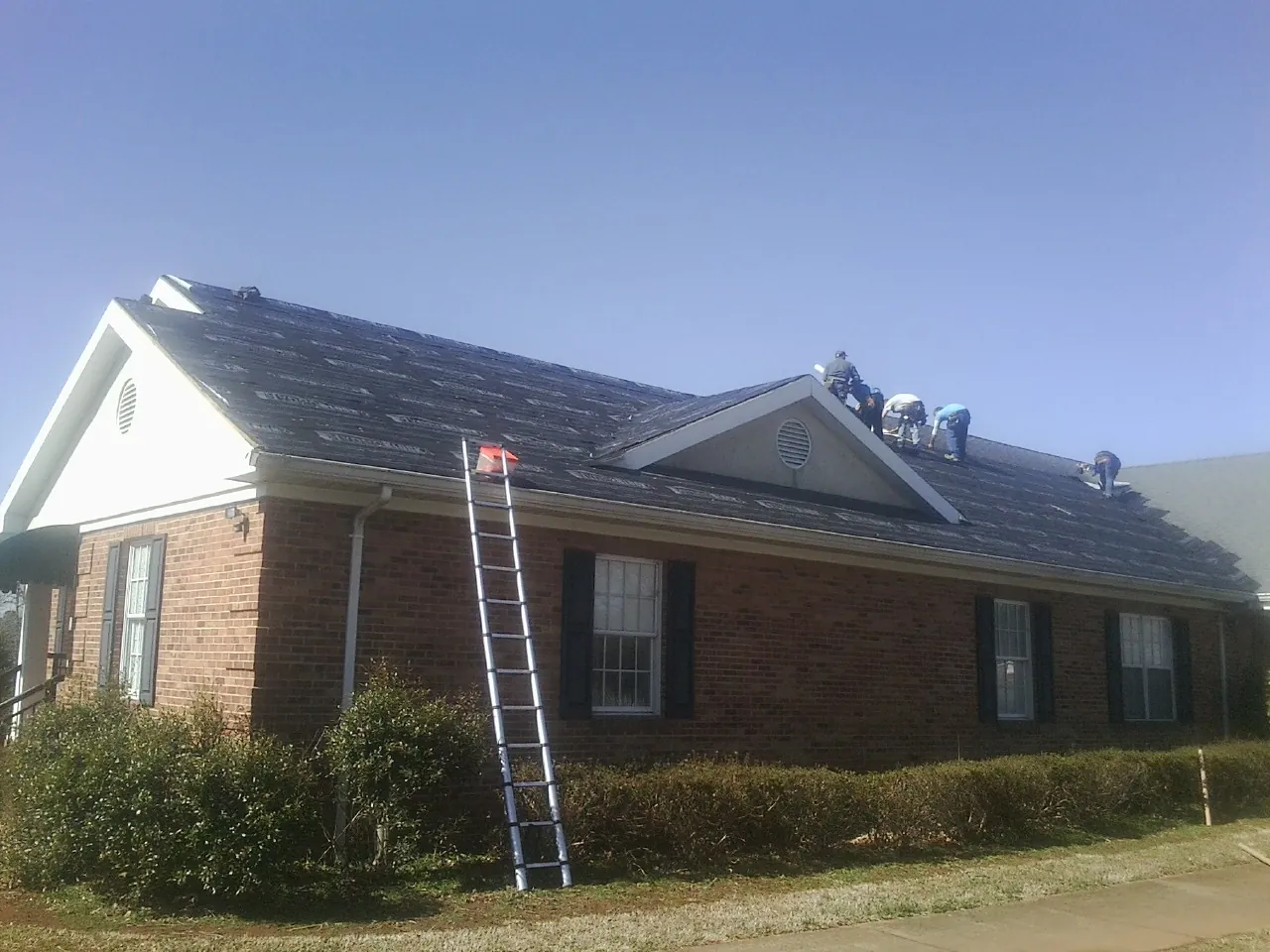 Crew tearing shingles off a brick ranch roof with a ladder set — Metal Roof Repair work by our crew in Lydia