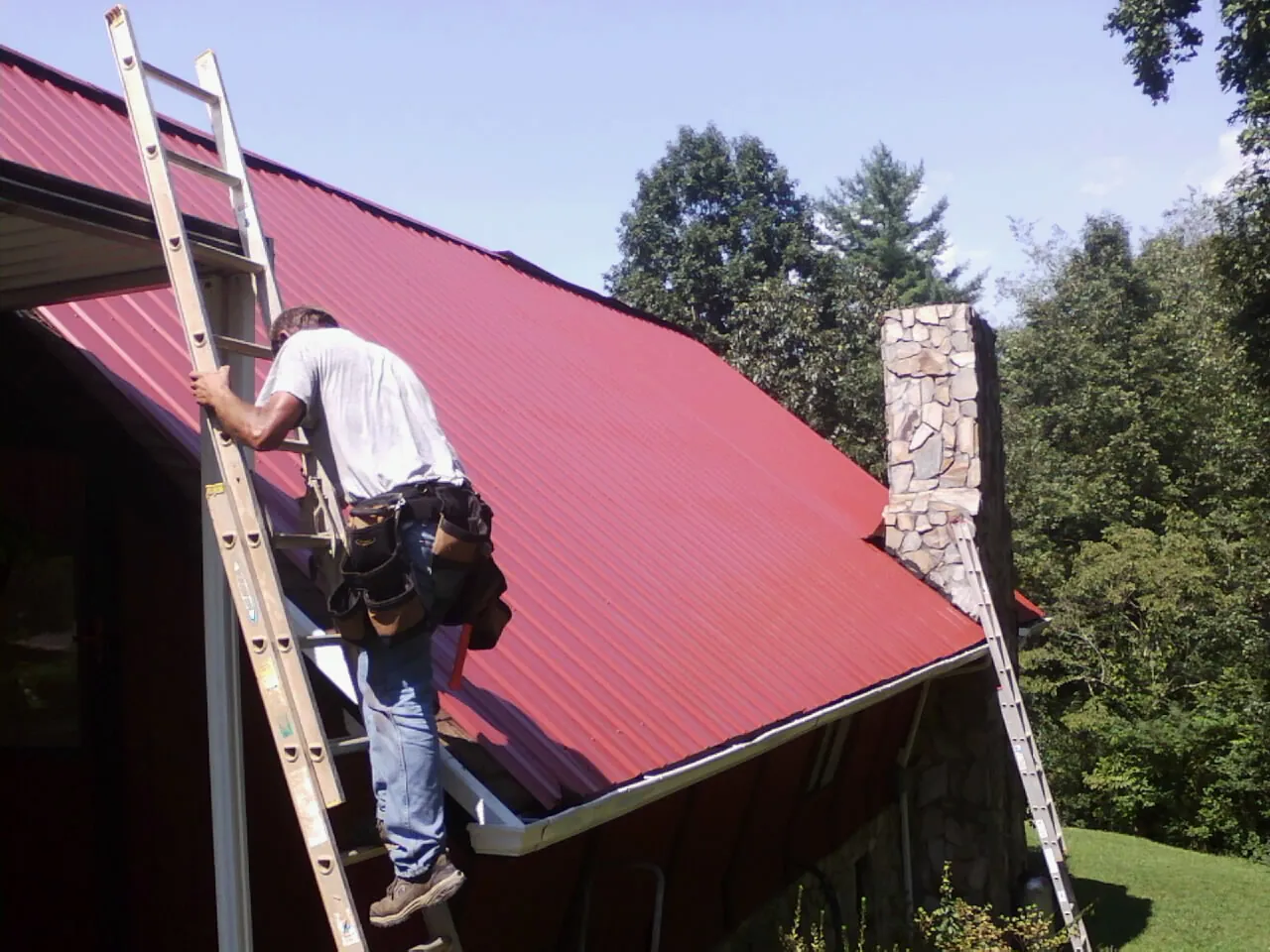 Roofer on a ladder beside a red metal roof and stone chimney — Soffit & Fascia Repair by Roofing Brothers in Lydia